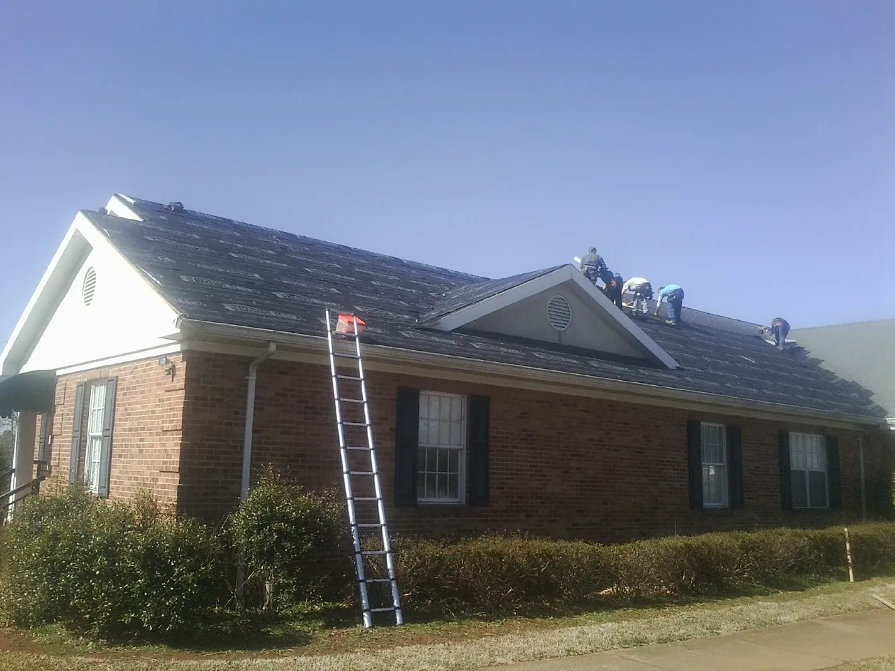 Crew tearing shingles off a brick ranch roof with a ladder set — Emergency Roof Repair work by our crew in Elrod