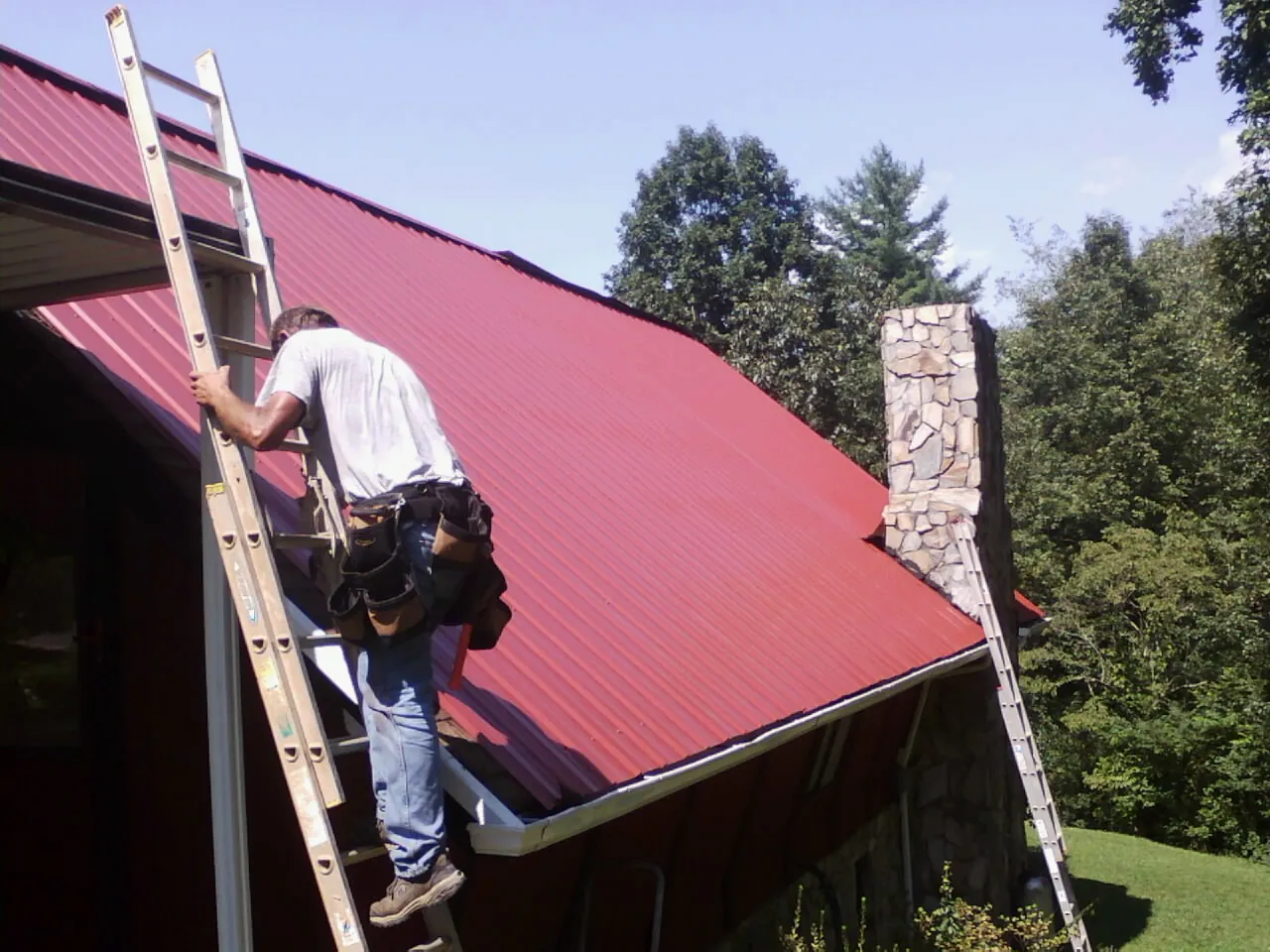 Roofer on a ladder beside a red metal roof and stone chimney — Soffit & Fascia Repair by Roofing Brothers in Elrod